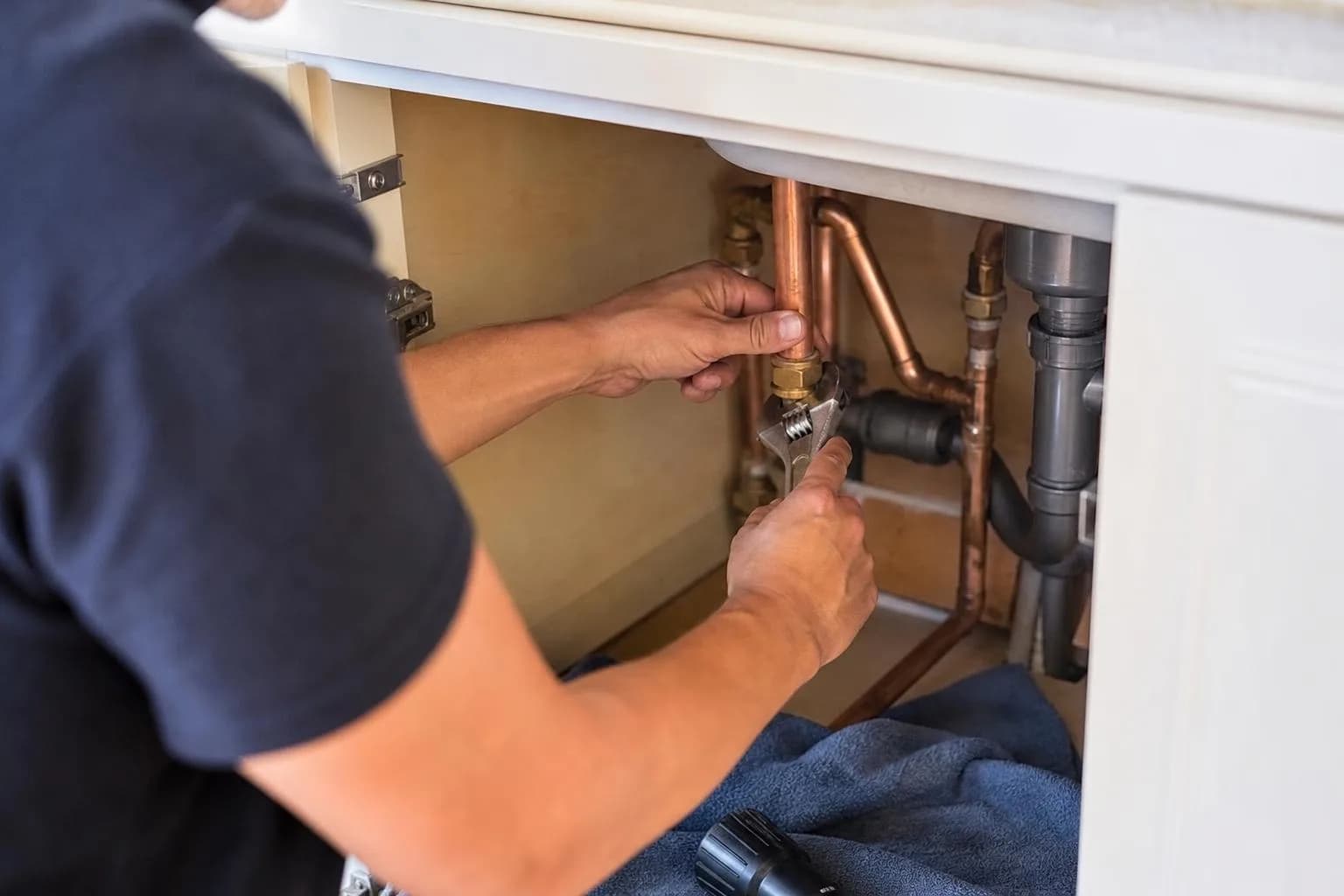 A plumber's hands tightening a compression joint on copper pipe under a kitchen sink